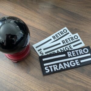 A photograph of a 3-inch orb on a 1-inch stand, with 2 black on white RetroStrange logo stickers and 1 white on black RetroStrange logo patch.
