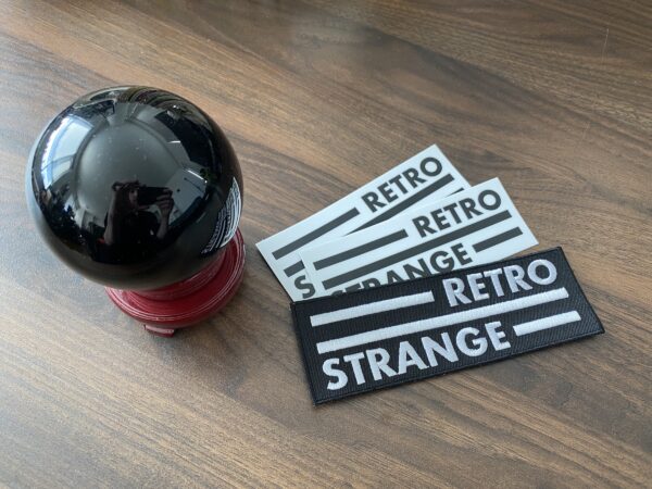 A photograph of a 3-inch orb on a 1-inch stand, with 2 black on white RetroStrange logo stickers and 1 white on black RetroStrange logo patch.
